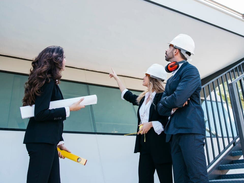 Three people in hard hats reviewing plans while inspecting a building exterior