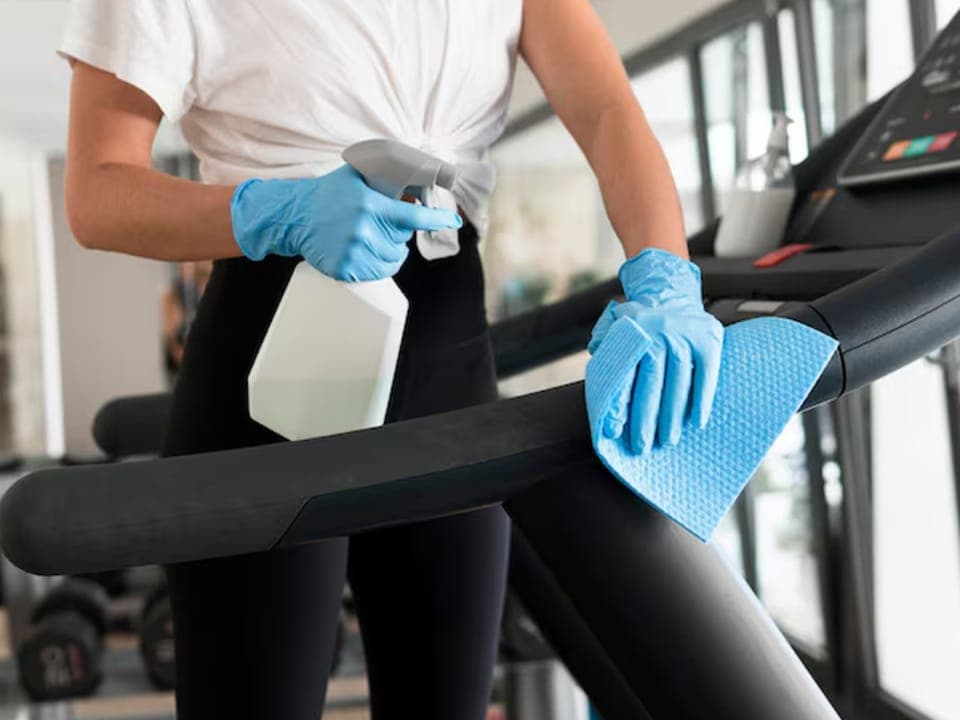 Gloved hands spraying and wiping down a treadmill handrail in a gym