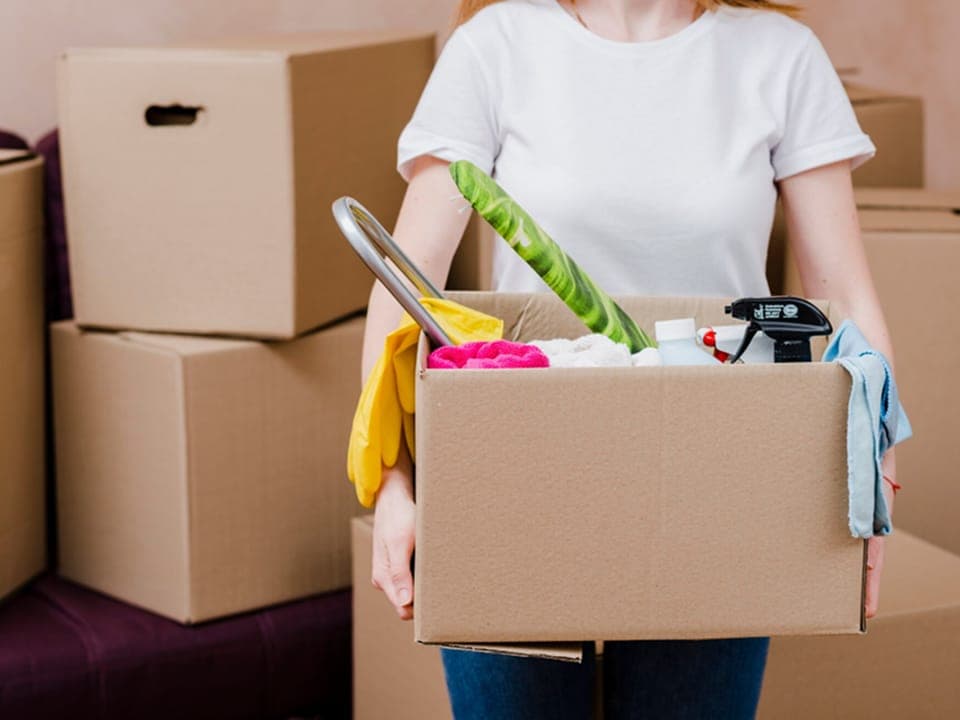 A cleaner carrying a box of supplies past stacked moving cartons