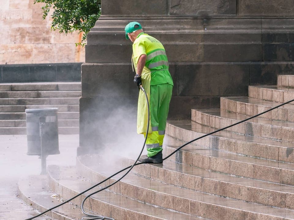 A worker in hi-vis pressure washing stone steps outside a building, spray misting off the stonework