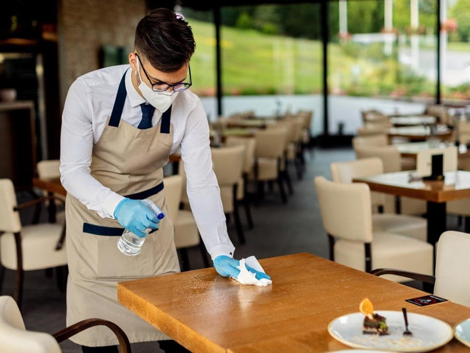 A masked, gloved staff member sanitising a table in an empty restaurant dining room